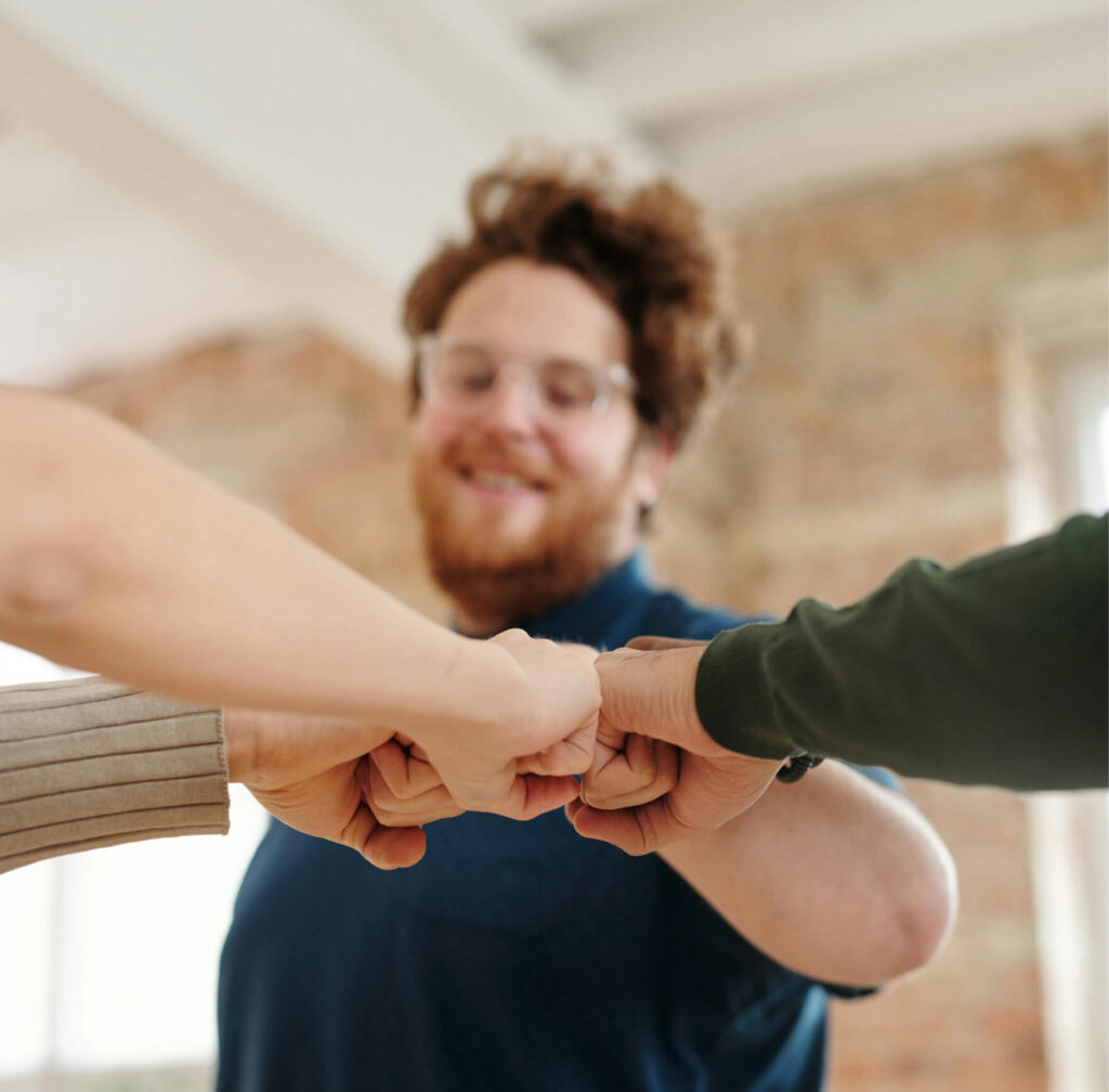 Vier Menschen geben sich in Teammanier einen Fauststoß. Einer von ihnen, ist unscharf zu erkennen, lächelt und blickt auf die zusammentreffenden Fäuste.