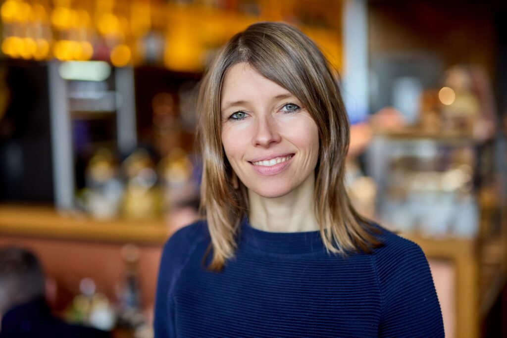 Anna Stolz lächelnd, sie trägt einen blauen Pullover. Sie hat schulterlange braune Haare.